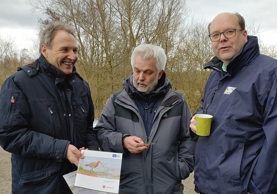 Three men stand next to each other. The man on the left presents a small booklet that features a Blak-tailed Godwit on the front. The man in the middle looks at the booklet. The man on the right smiles into the camera.